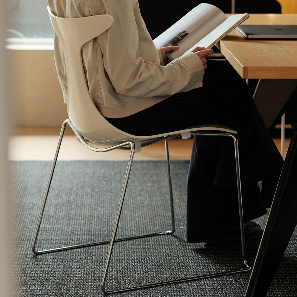 A person seated on a modern white molded chair with a slim metal frame, reading an open book at a wooden desk in a bright indoor setting.