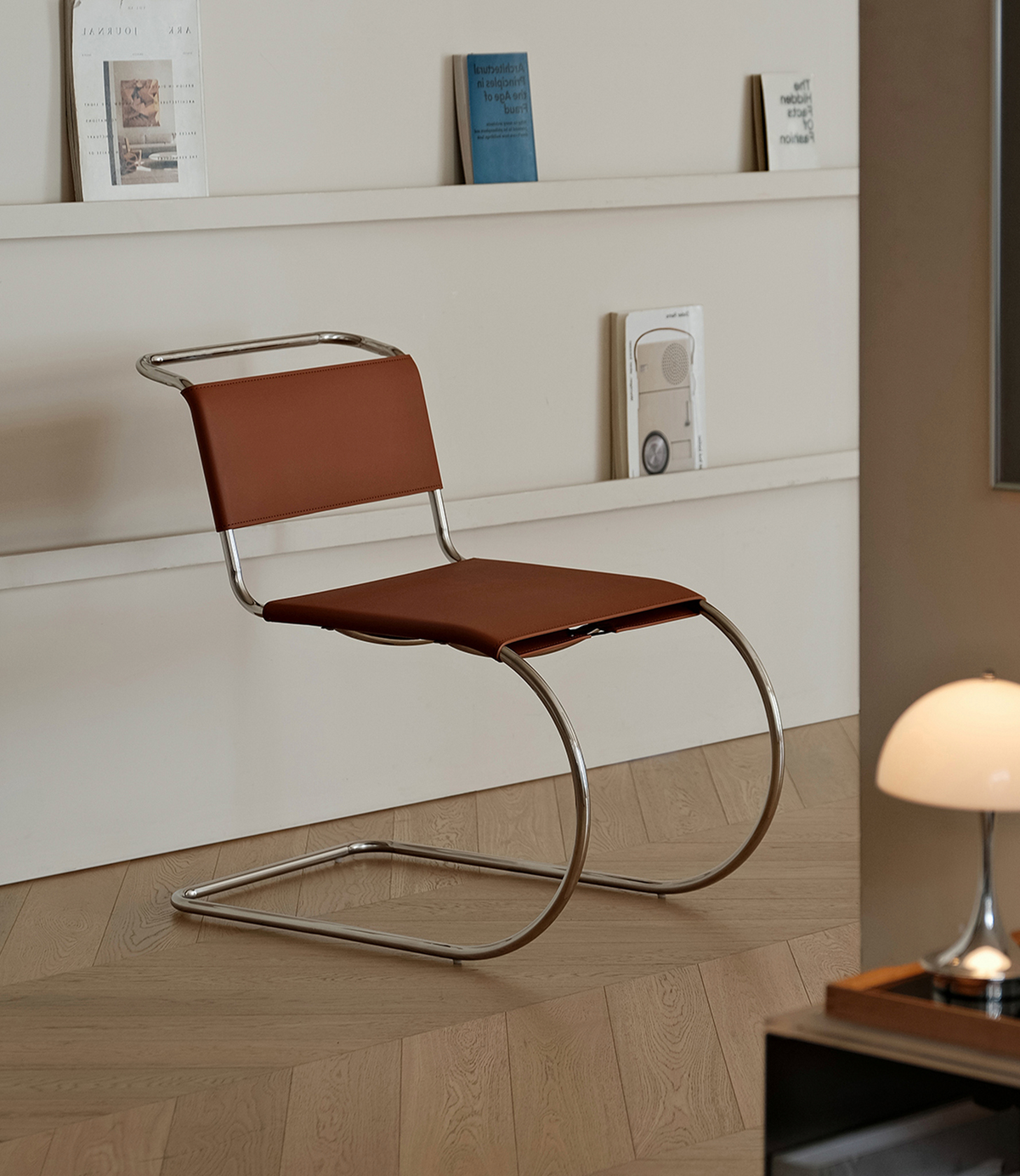A modern cantilever chair with a brown upholstered seat and backrest and a curved chrome metal frame, shown in a minimalist interior setting with a wooden floor, white wall shelves, and a lamp.