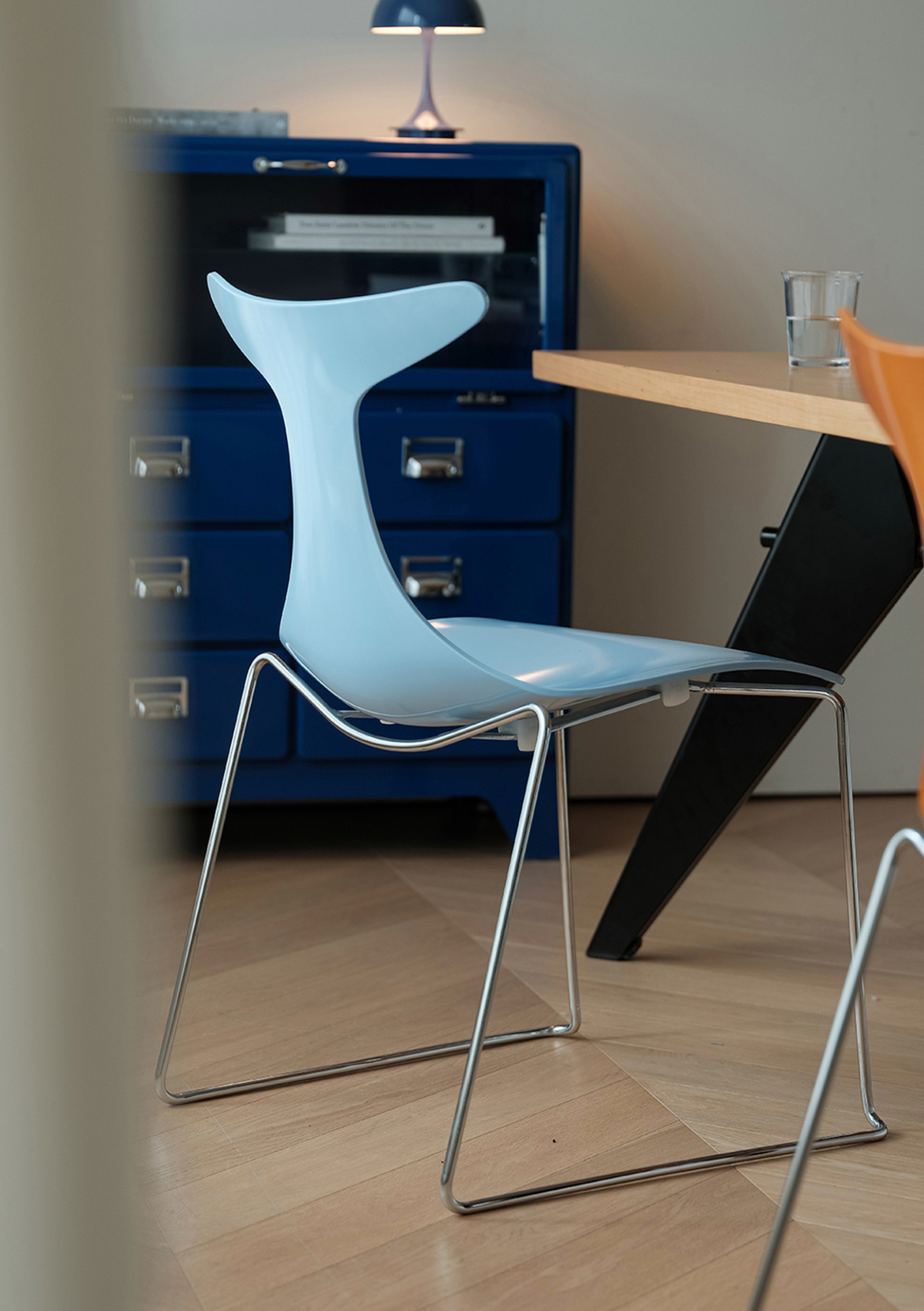 A light blue molded plastic chair with a chrome metal wire frame, positioned in a dining or office setting next to a wooden table and a dark blue cabinet.