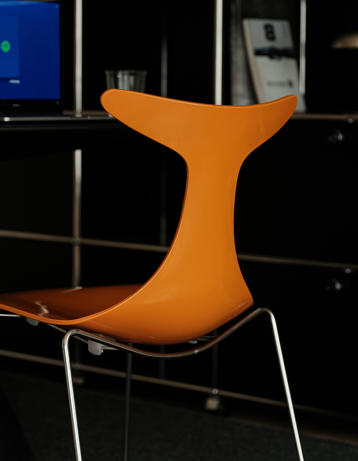 An orange molded plastic chair with a chrome metal wire frame, shown in a modern office setting with a dark desk and computer monitor in the background.