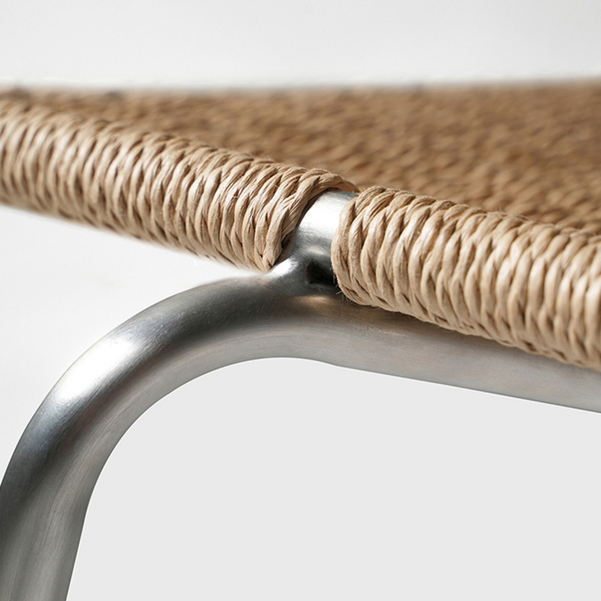 Close-up of a woven natural-fiber seat strap attached to a curved silver metal frame, shown against a light gray background.