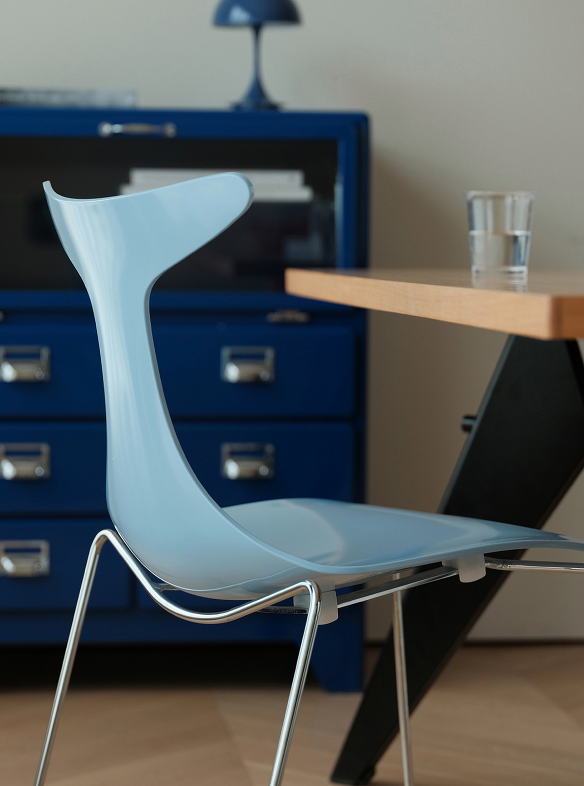A light blue molded plastic chair with a curved back and seat, supported by thin silver metal legs, positioned next to a wooden table in a modern room setting.