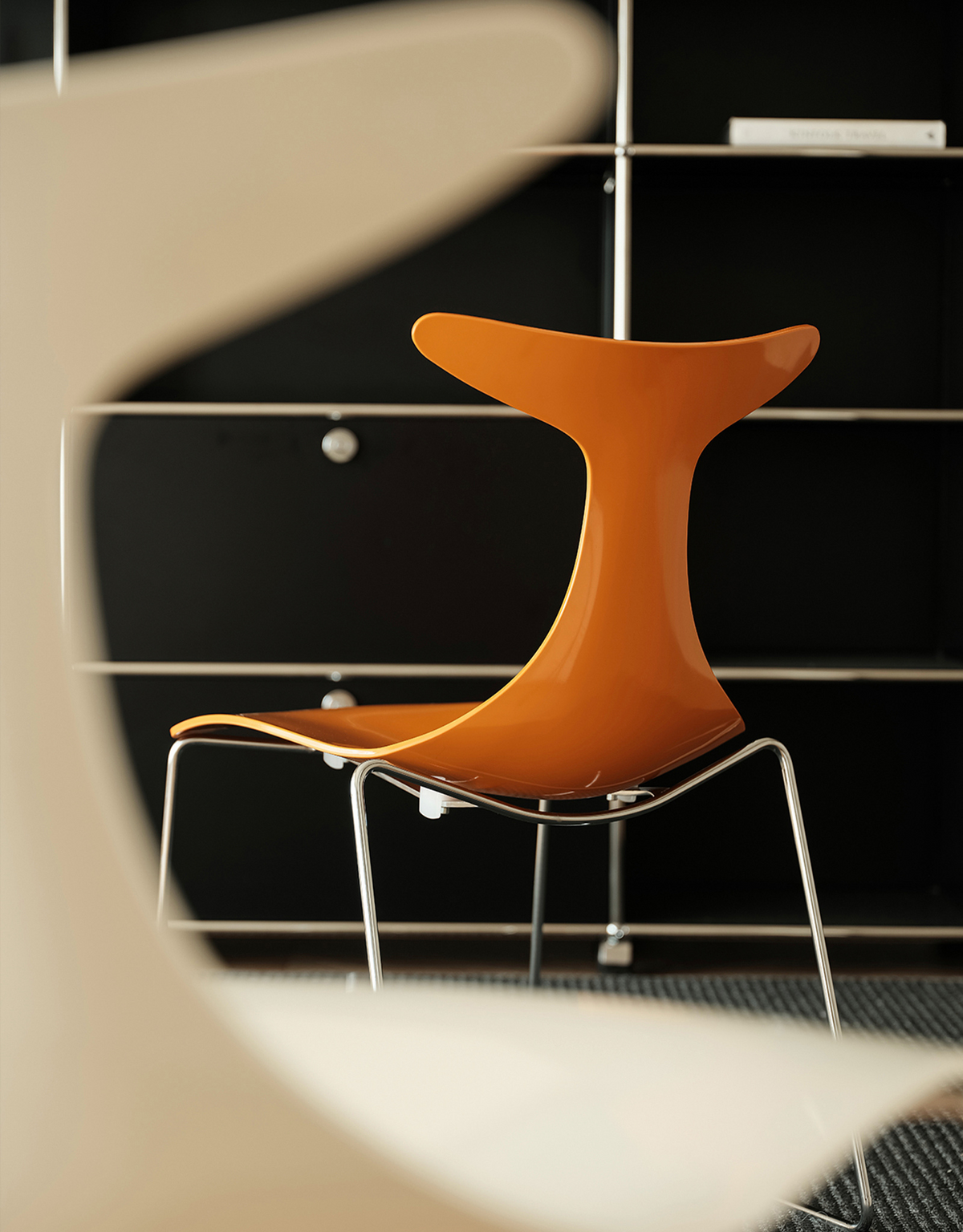 An orange molded plastic chair with a chrome wire frame, shown from a low angle with a blurred foreground element.