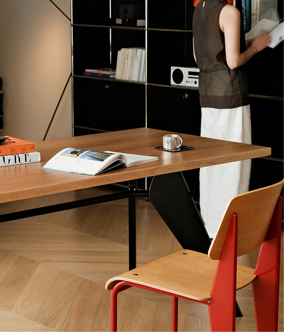 A modern wooden desk in a minimalist room setting, with an open book, a coffee cup, and a person standing in the background near black shelving. A red and light wood chair is prominently featured in the foreground.