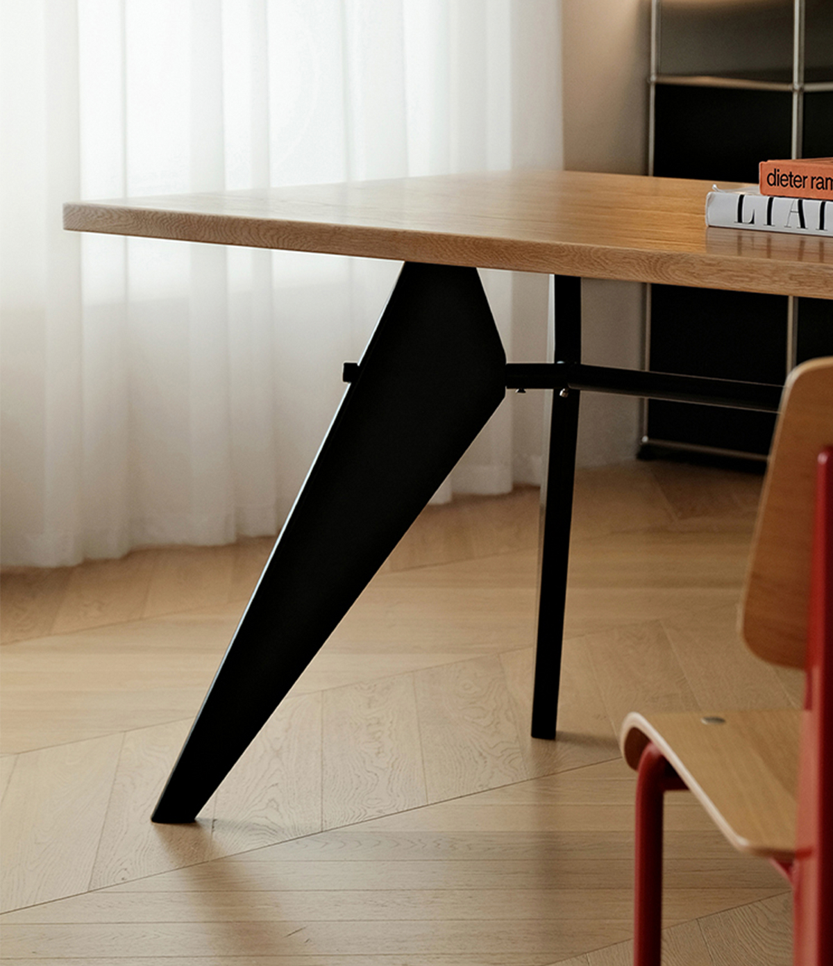 A modern writing desk with a light wood top and a bold black angled metal base, shown in a bright room with light wood flooring and a matching chair partially visible.