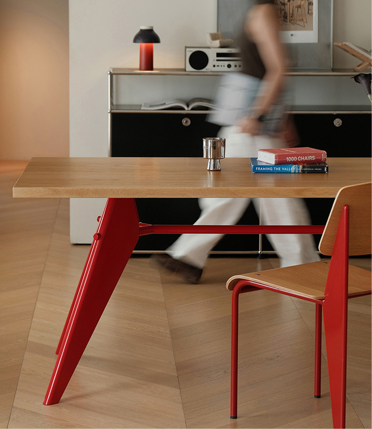 A modern dining table with a light wood top and bold red metal legs, paired with a matching red and wood chair in a contemporary interior. A blurred person walks in the background near a cabinet.