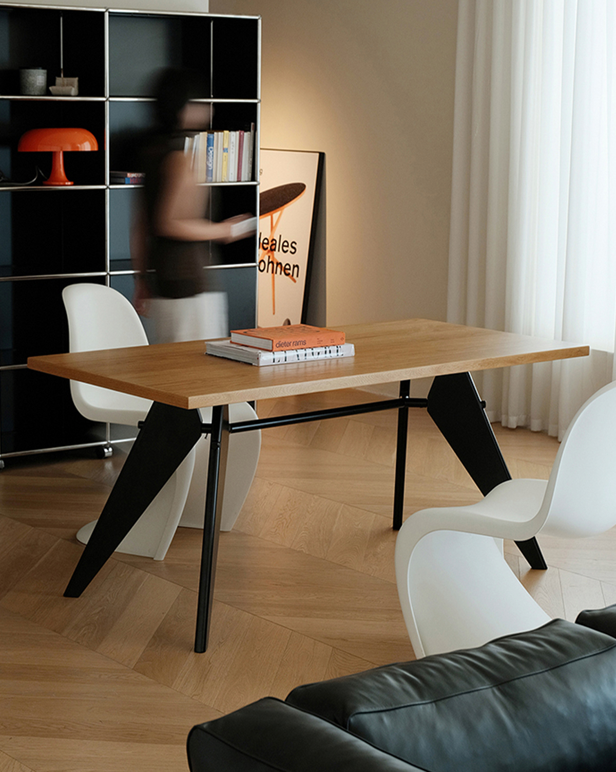A rectangular wooden dining table with black angled metal legs, styled in a modern room with white chairs, a bookshelf, and a light wood floor.