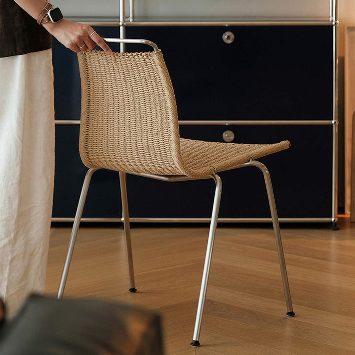 A woven natural-fiber chair with a curved seat and backrest, supported by slim silver metal legs, shown in a room setting with a person's hand resting on the backrest.