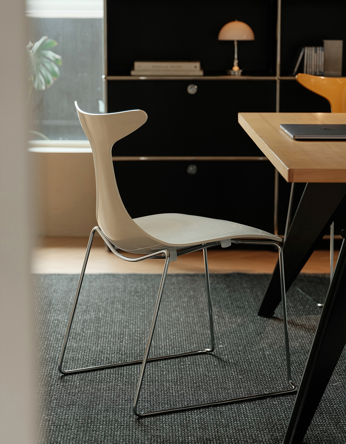 A modern light beige molded plastic chair with a chrome wire frame, positioned next to a wooden desk in a contemporary home office setting.