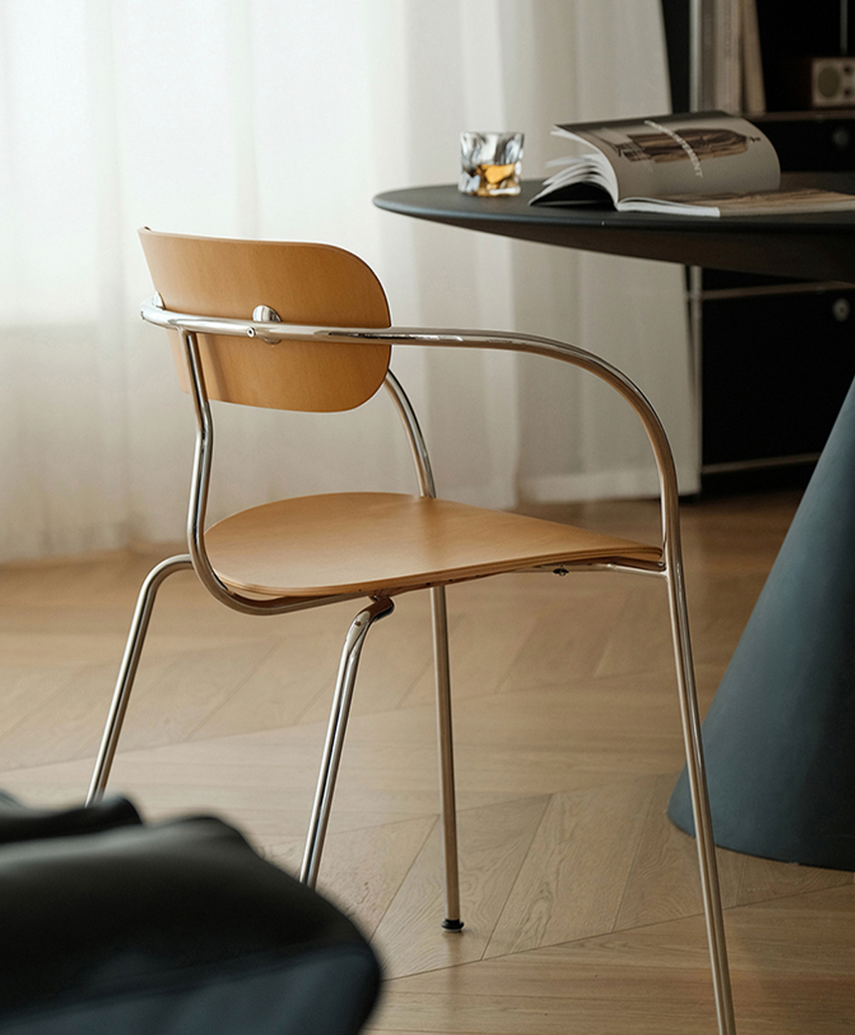 A modern wooden dining chair with a curved chrome metal frame, shown in a bright, contemporary room next to a dark table.