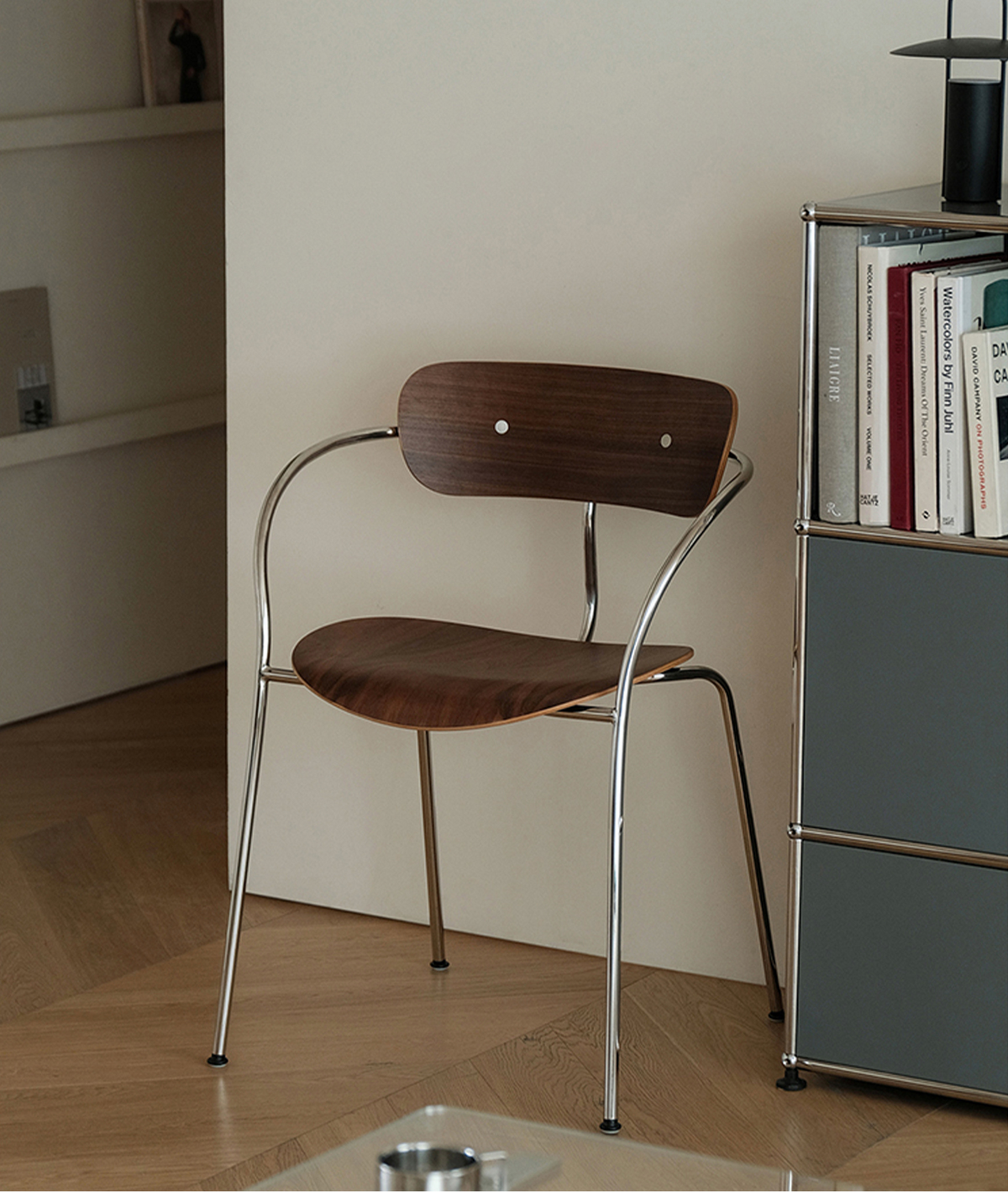 A mid-century modern wooden dining chair with a curved chrome metal frame, positioned next to a gray bookshelf in a minimalist room.