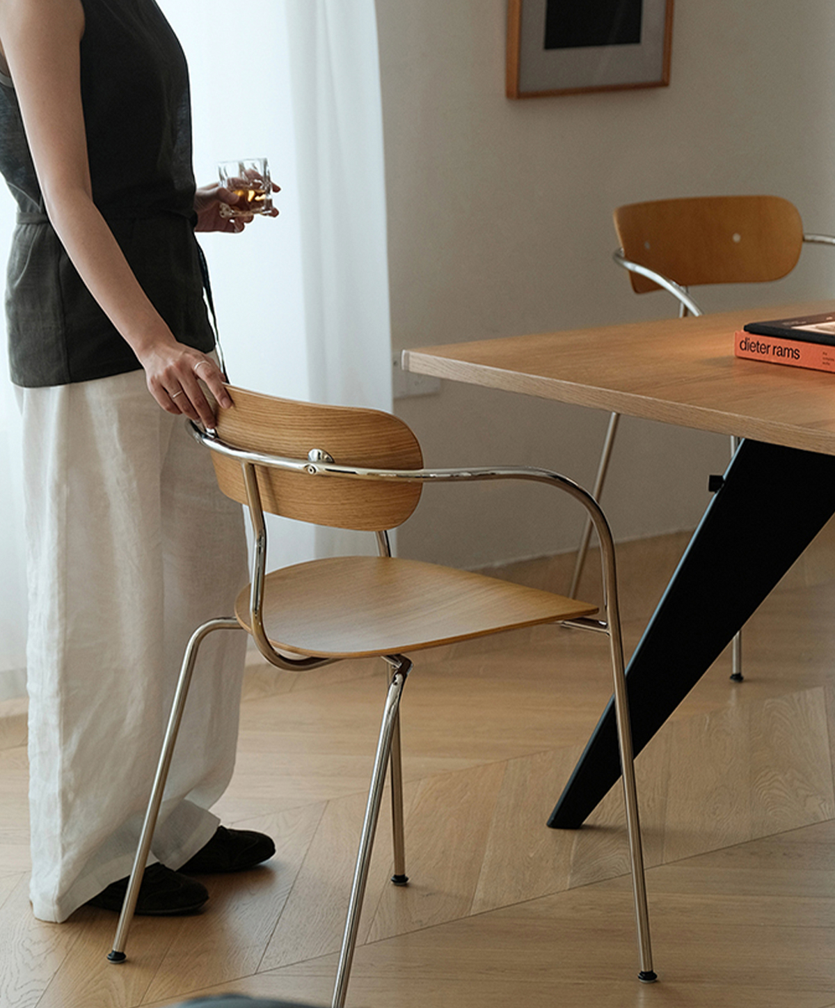 A modern wooden dining chair with a curved chrome metal frame and seat, shown in a dining room setting next to a wooden table. A person is partially visible on the left holding the chair's backrest.