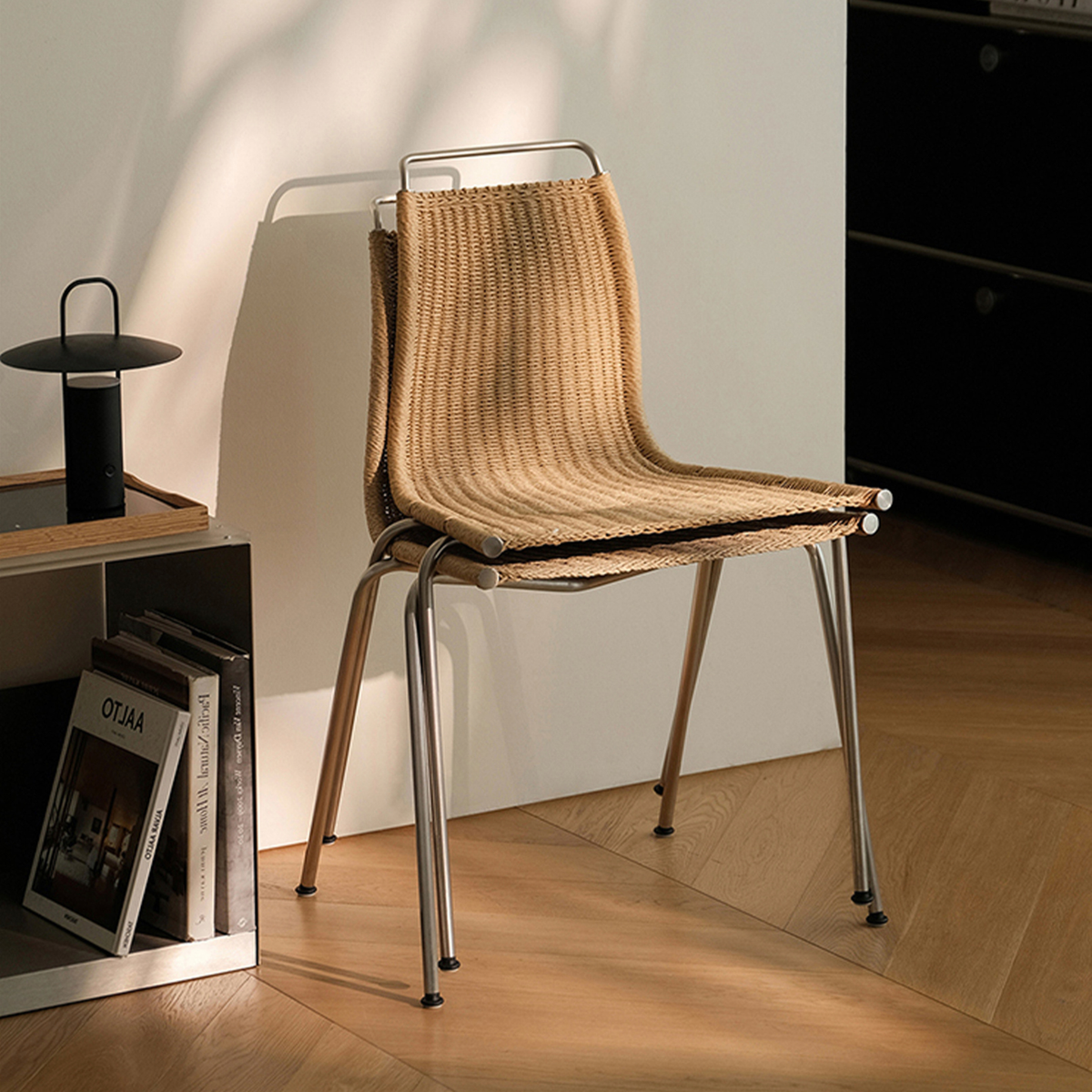 A modern woven chair with a light brown textured seat and backrest, supported by a chrome metal frame, set in a sunlit room with a wooden floor and a white wall.