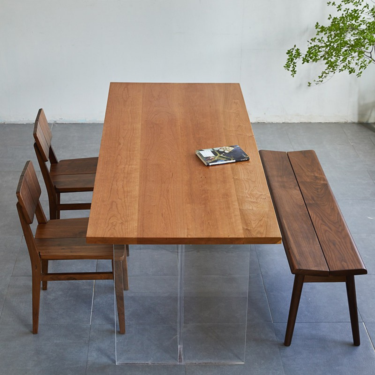 A rectangular wooden dining table with a warm medium-brown finish, paired with two matching wooden chairs and a long wooden bench. The table is supported by clear acrylic legs, and a magazine rests on the tabletop.