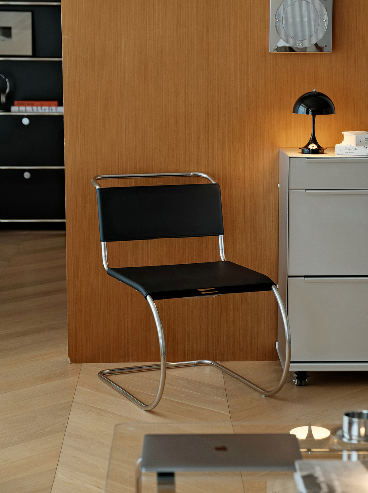 A modern cantilever chair with a black seat and backrest and a chrome metal frame, positioned in a contemporary office or living space next to a light gray cabinet.