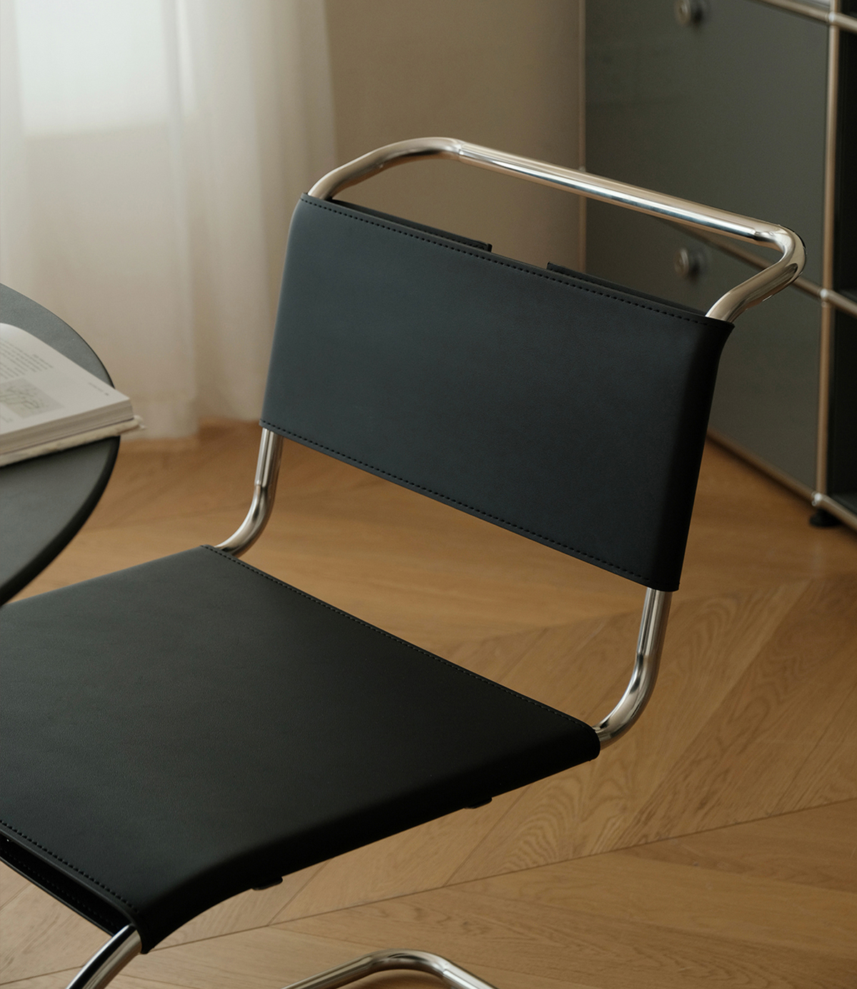 A modern black upholstered chair with a curved chrome metal frame, shown in a room setting with a light wood floor and a dark cabinet in the background.