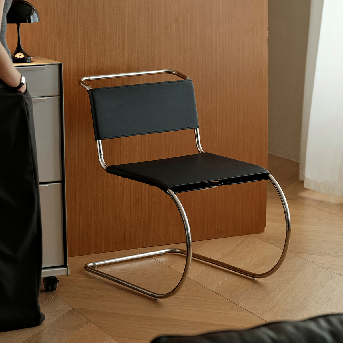 A modern cantilever dining chair with a black upholstered seat and backrest and a curved chrome metal base, shown in a contemporary room setting with a wooden wall and light wood floor.
