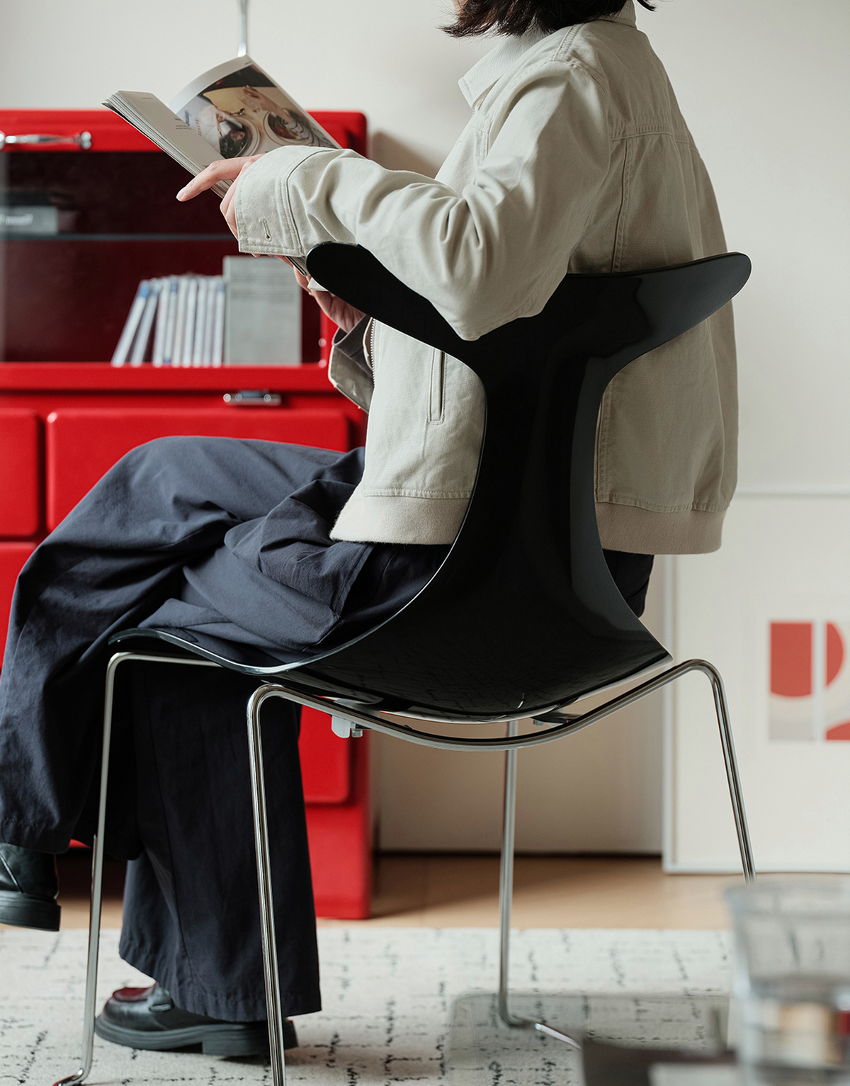 A person seated on a glossy black molded chair with chrome legs, reading an open book in a modern interior with a red cabinet in the background.