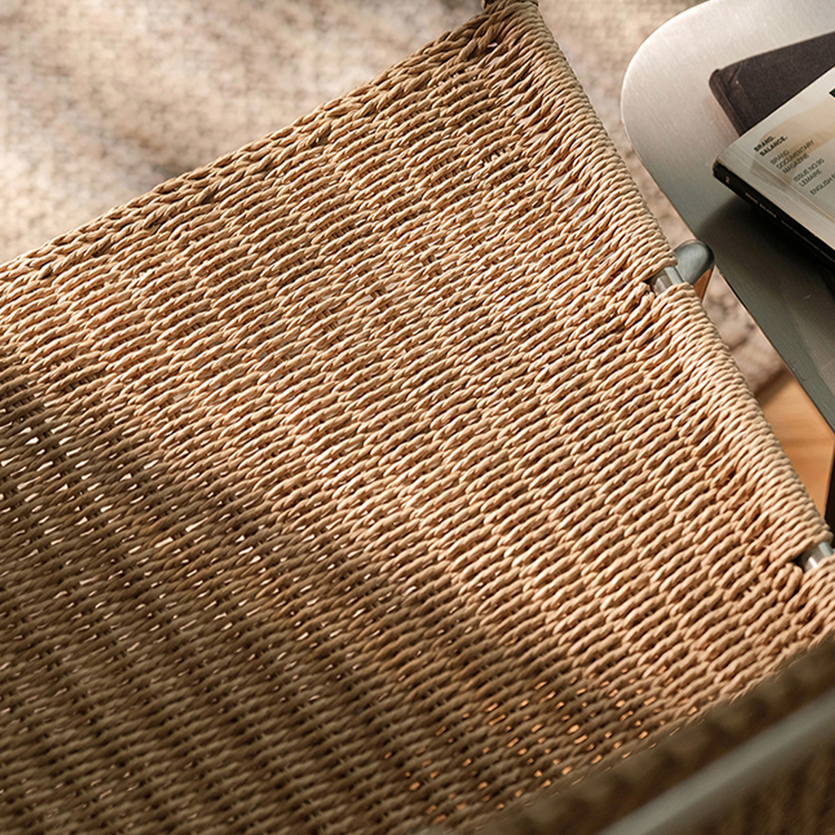 Close-up of a woven natural-fiber chair or bench seat in a warm tan color, with part of a white chair and a book visible in the background.