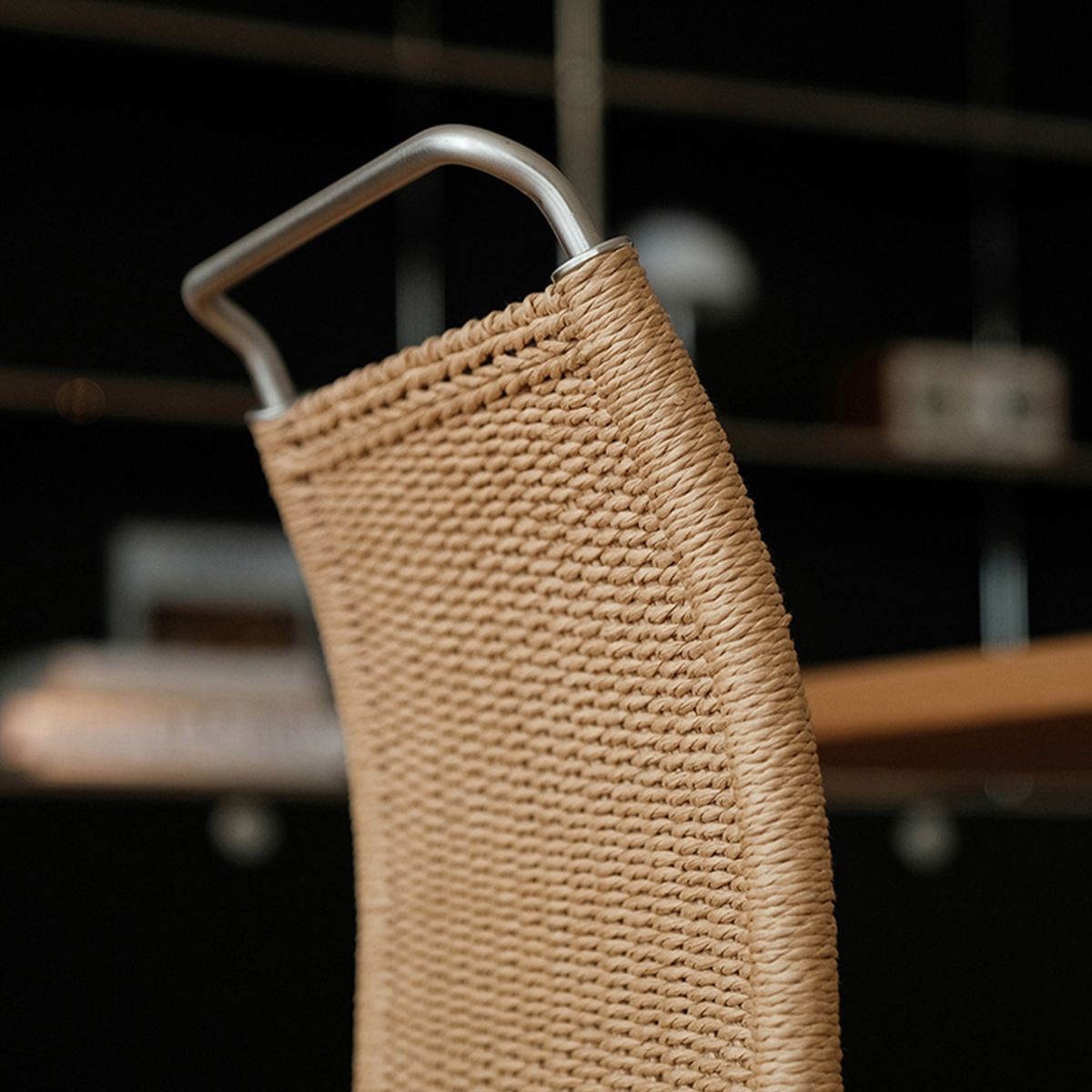 Close-up of a woven rattan or rope chair back in a natural tan color, attached to a curved silver metal frame, with a dark blurred interior background.