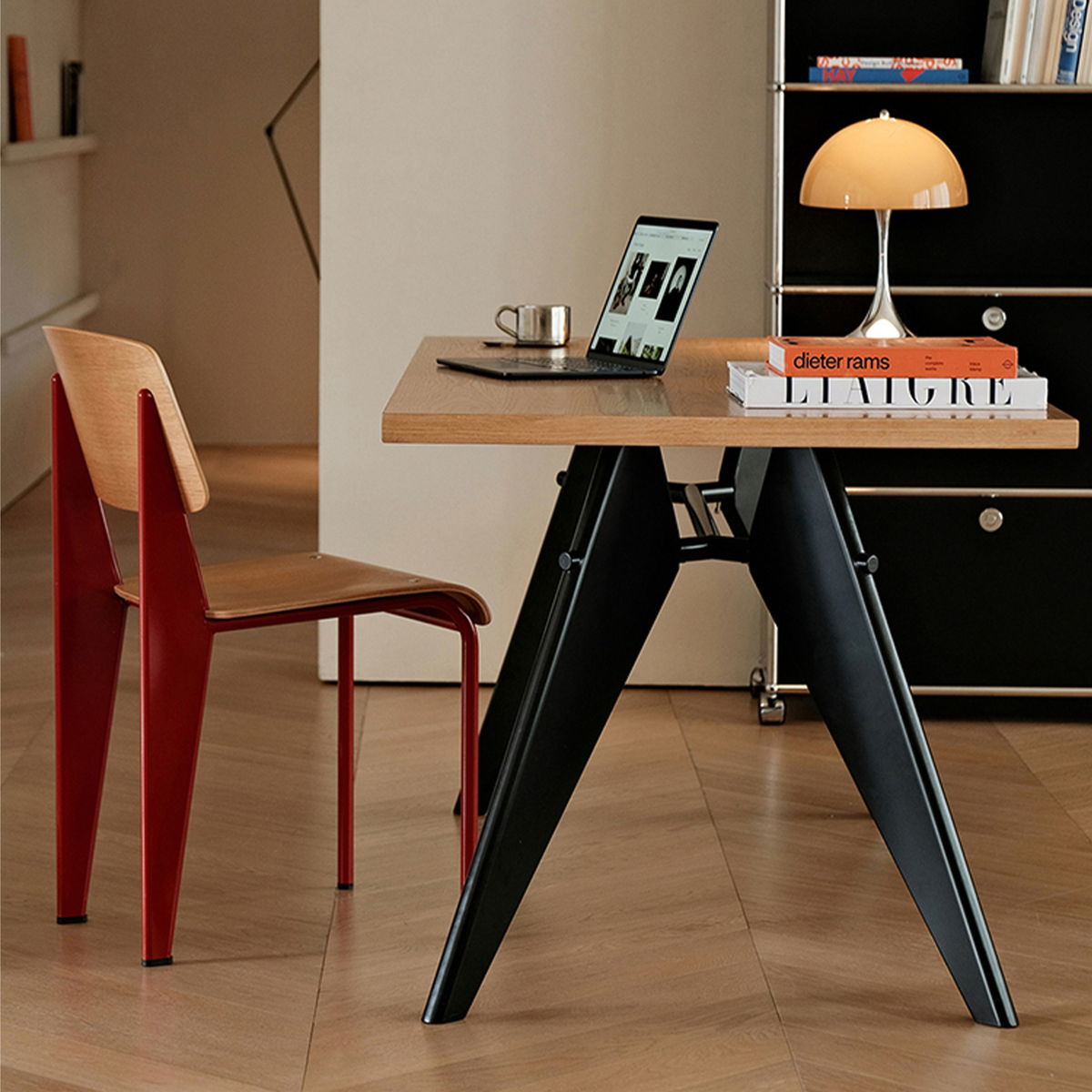 A modern writing desk with a light wood top and black angled metal legs, styled in a home office setting with a red chair, laptop, and lamp.