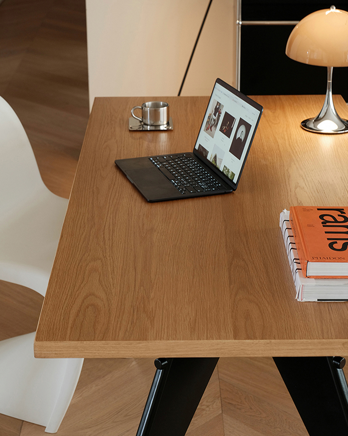 A light brown wood-grain desk with black metal legs, styled with an open laptop, a white chair, a lamp, and books on a light-colored floor.