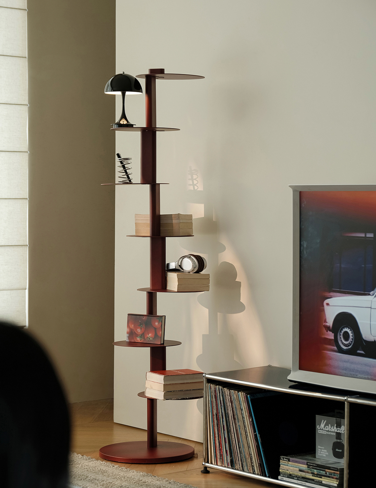 A tall, narrow wooden shelving unit with multiple tiers and a round base, styled in a living room next to a TV stand and television.