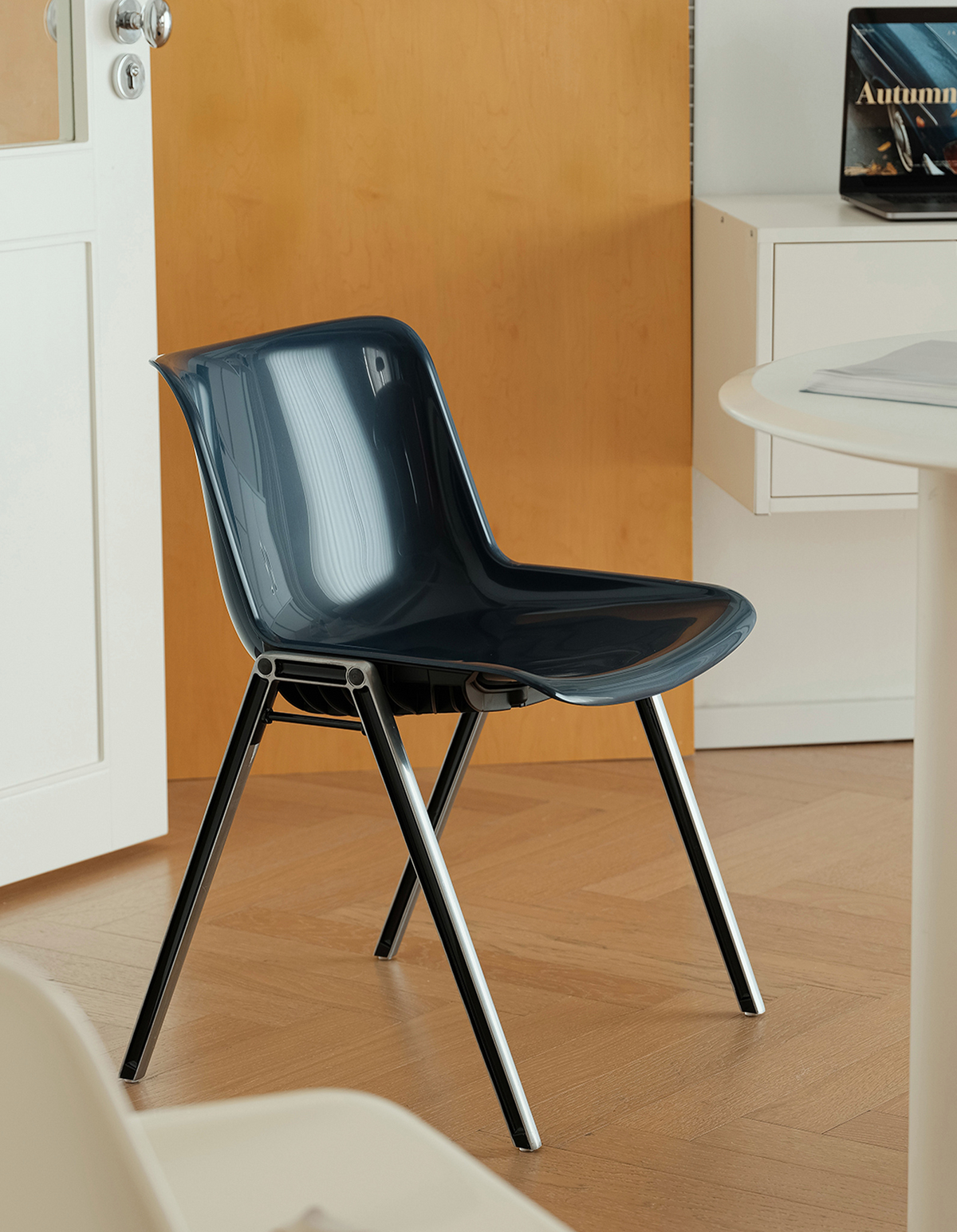 A modern dark blue-green molded plastic chair with black metal legs, shown in a home office setting with a wooden floor, white desk, and cabinet in the background.