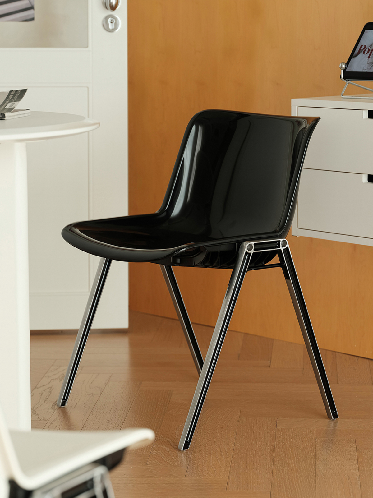 A glossy black molded plastic chair with slim black metal legs, shown in a modern room setting with a wood floor, white furniture, and a wooden wall panel.