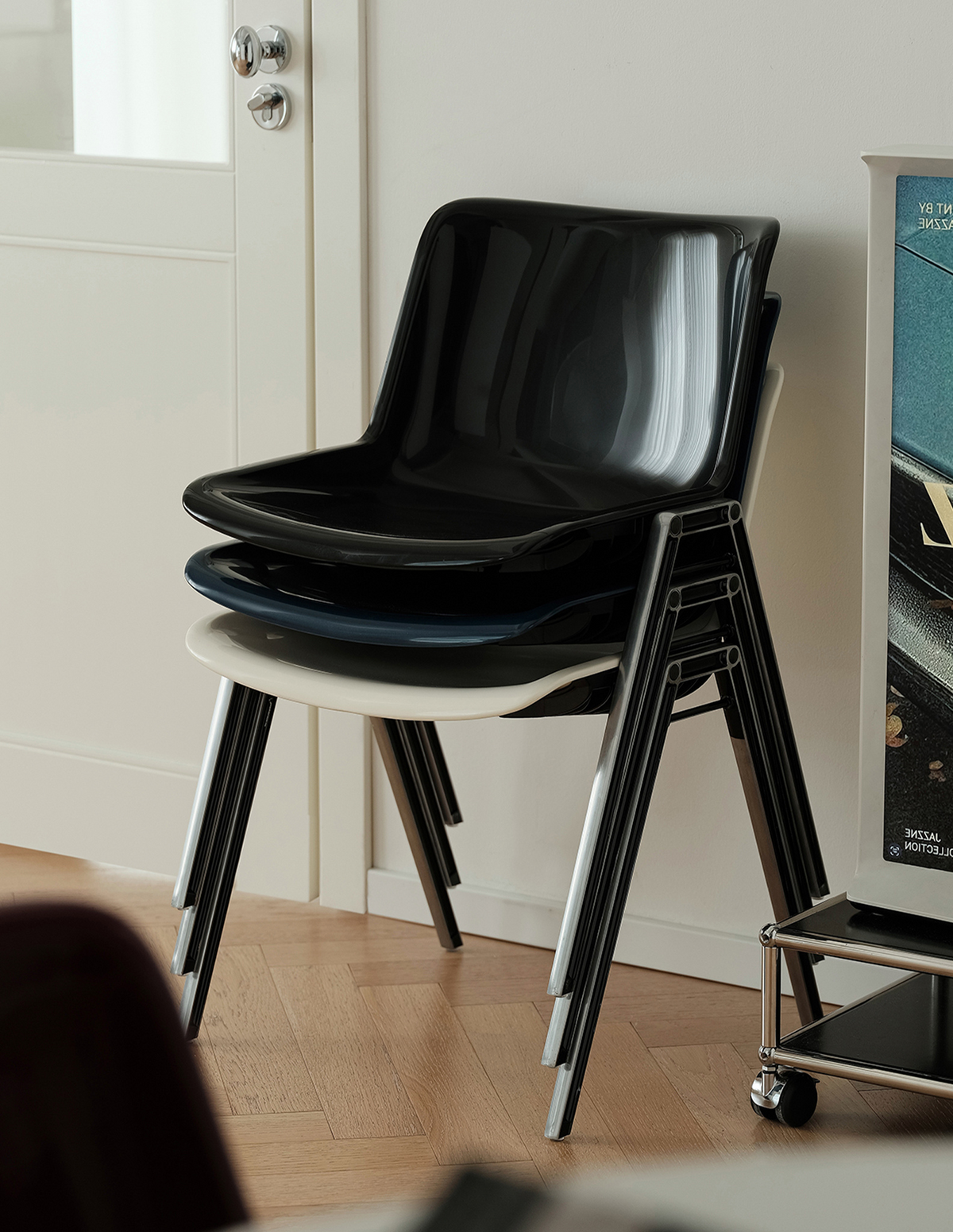A stack of glossy black molded plastic chairs with silver metal legs, shown in a home interior setting next to a white door and a poster.