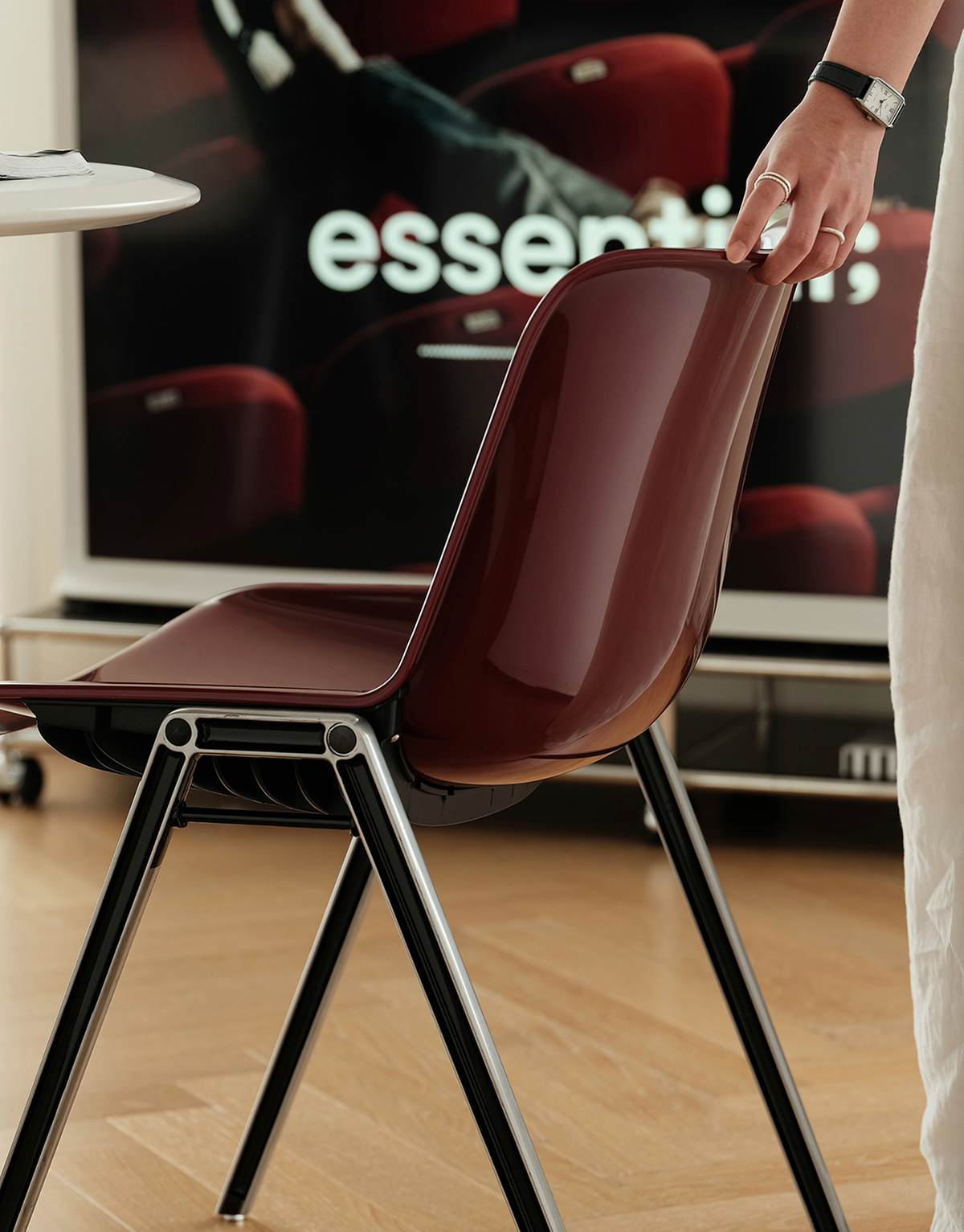 A glossy dark burgundy molded plastic chair with black metal legs, shown in a modern room setting with a person's hand resting on the backrest.