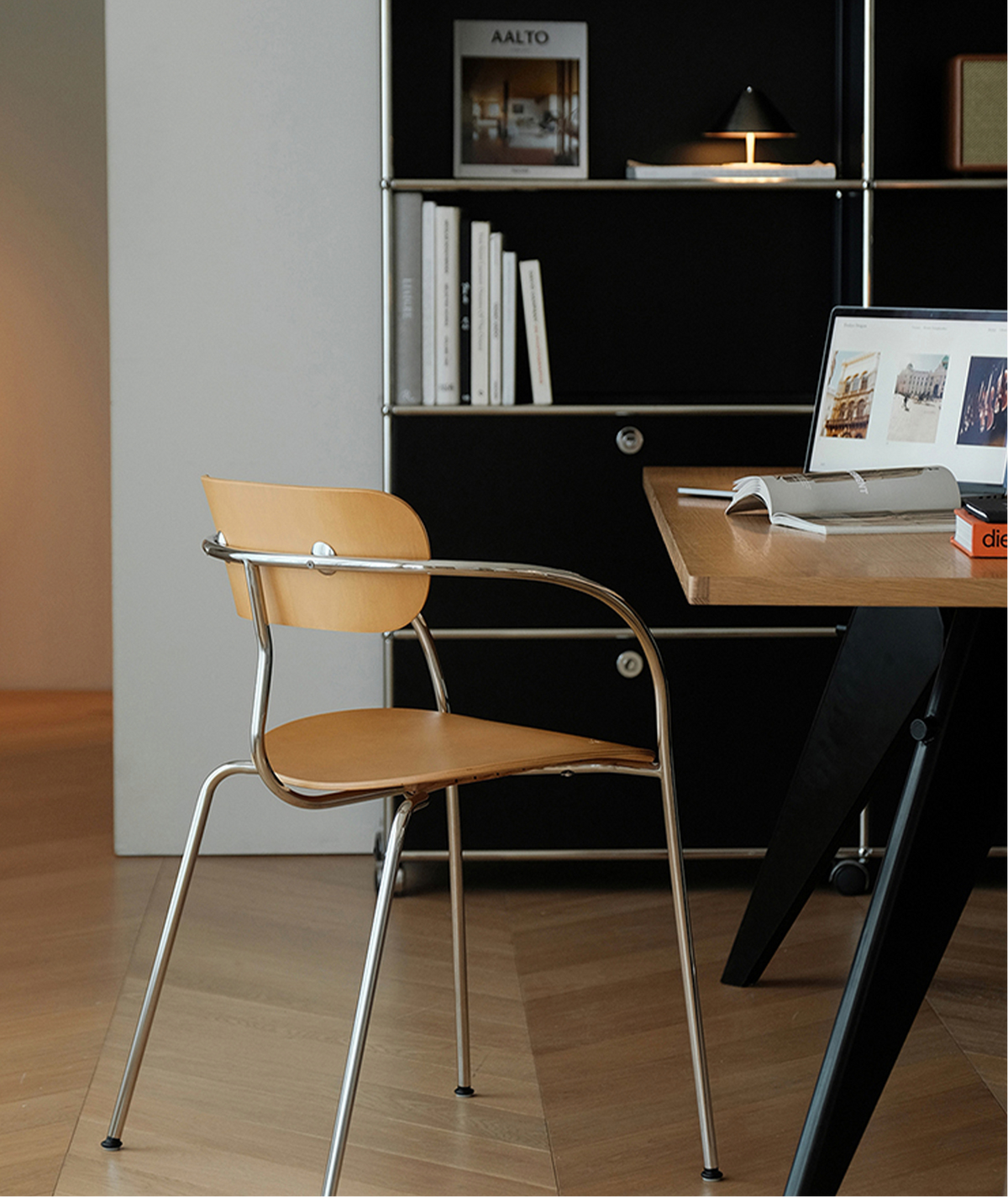 A modern wooden chair with a curved chrome metal frame sits beside a light wood desk in a minimalist room with a black shelving unit in the background.