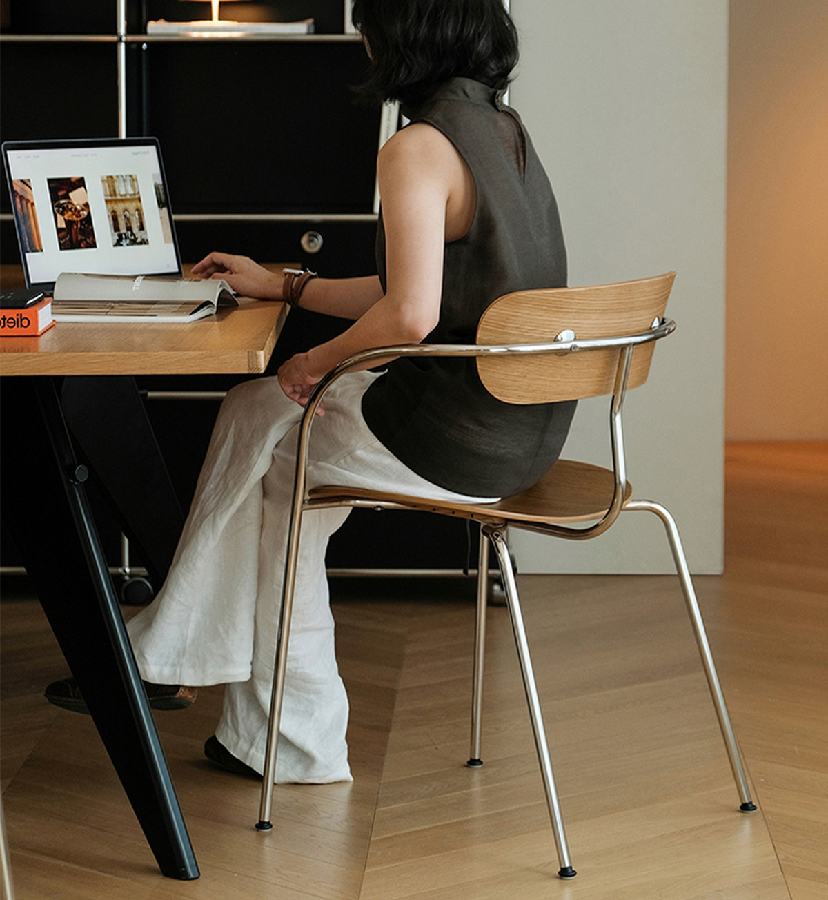 A woman sits on a modern wooden and metal chair at a desk, working on a laptop in a minimalist home office setting.