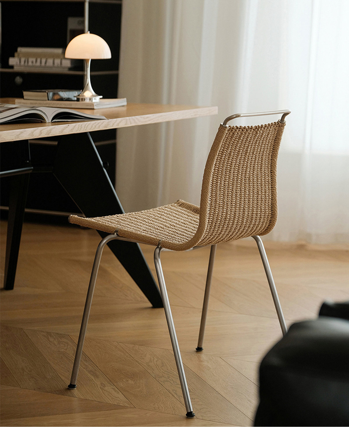 A modern woven rattan chair with a curved silver metal frame, shown in a bright, contemporary room with a wooden desk and lamp in the background.