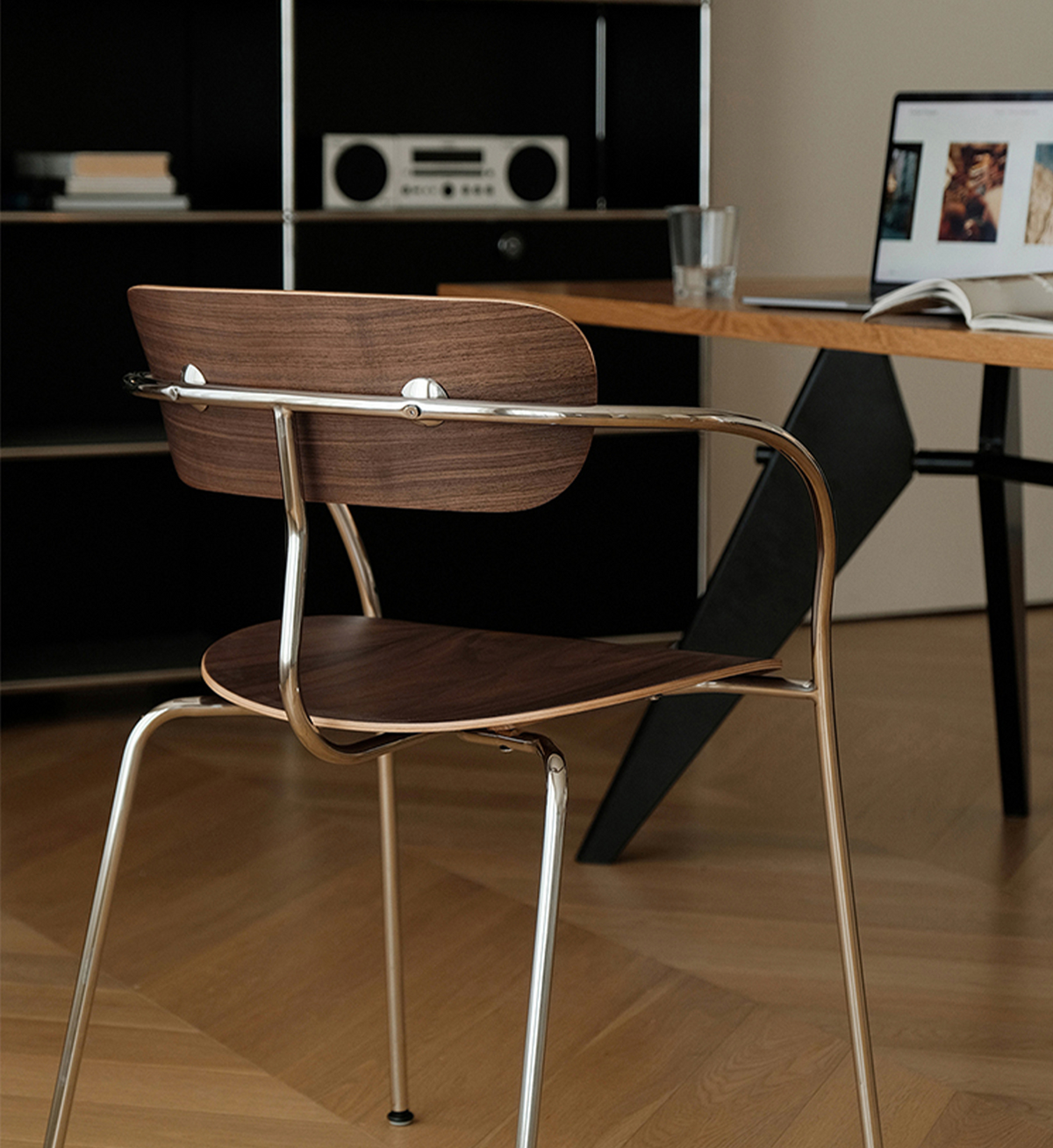 A modern wooden armchair with a curved chrome metal frame and seat, shown in a home office setting next to a desk with a laptop and glass.