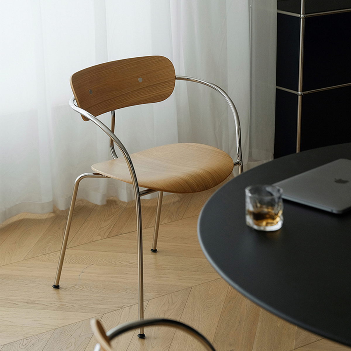 A modern wooden dining chair with a curved chrome metal frame, shown in a bright room next to a black round table with a laptop and a glass of whiskey.