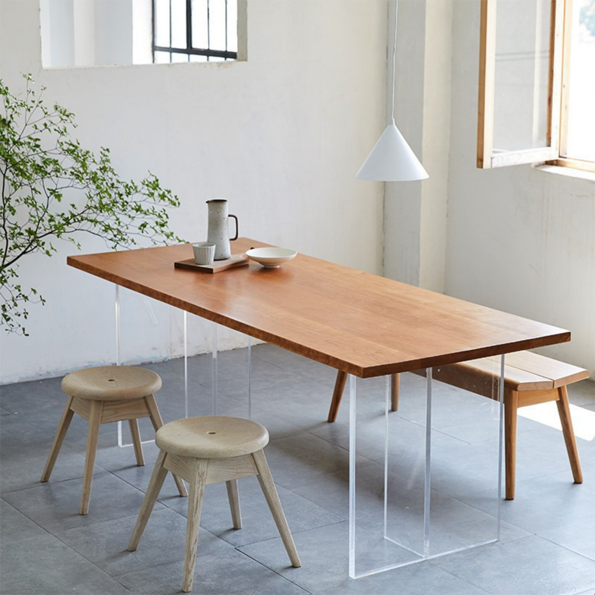 A rectangular wooden dining table with clear acrylic legs, shown in a bright minimalist room with two wooden stools, a pendant light, and a plant.