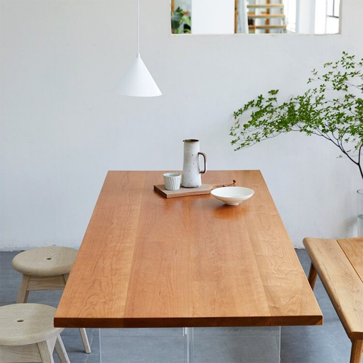 A rectangular wooden dining table with a warm honey finish, styled in a bright, minimalist room with a white wall, a hanging pendant light, and simple wooden stools.