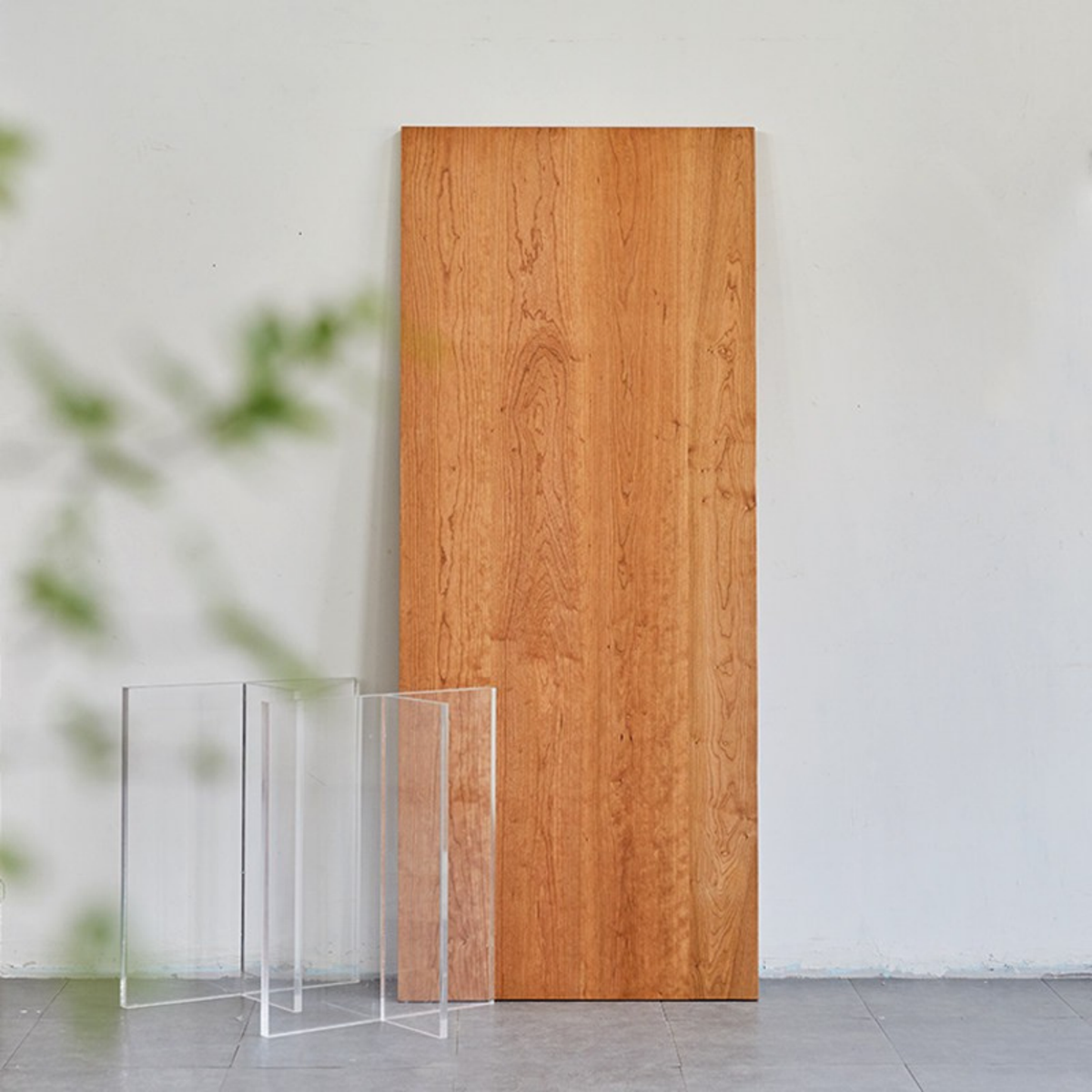 A tall rectangular wood panel with a warm honey-brown wood grain stands upright against a light gray wall, with three clear acrylic display blocks in front on a gray floor.