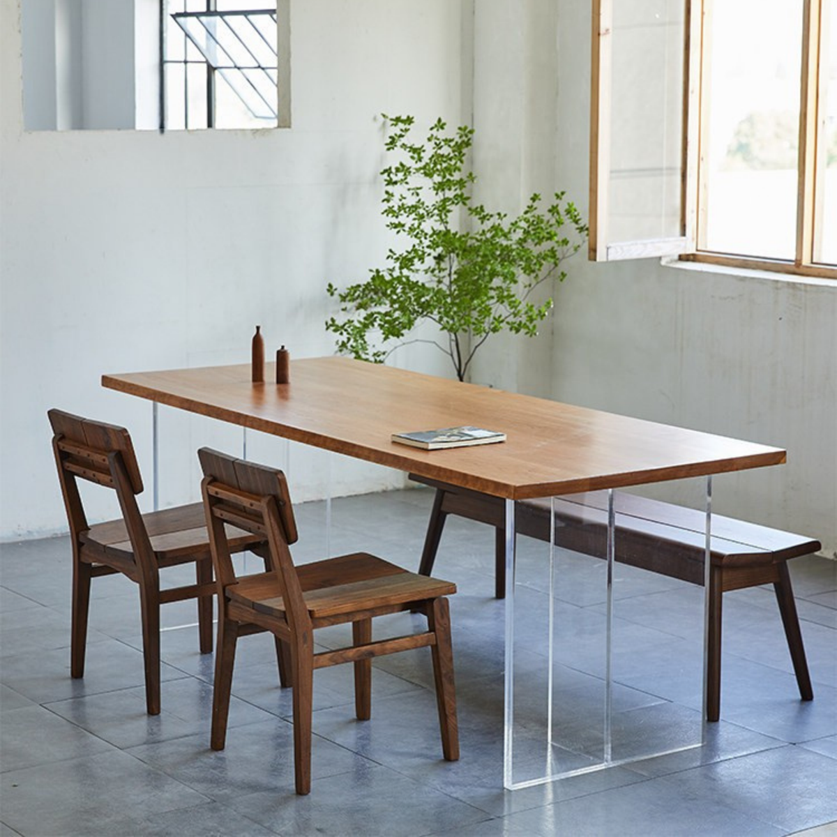 A rectangular wooden dining table with clear acrylic legs, accompanied by two wooden chairs and a matching wooden bench in a bright, modern room with large windows.